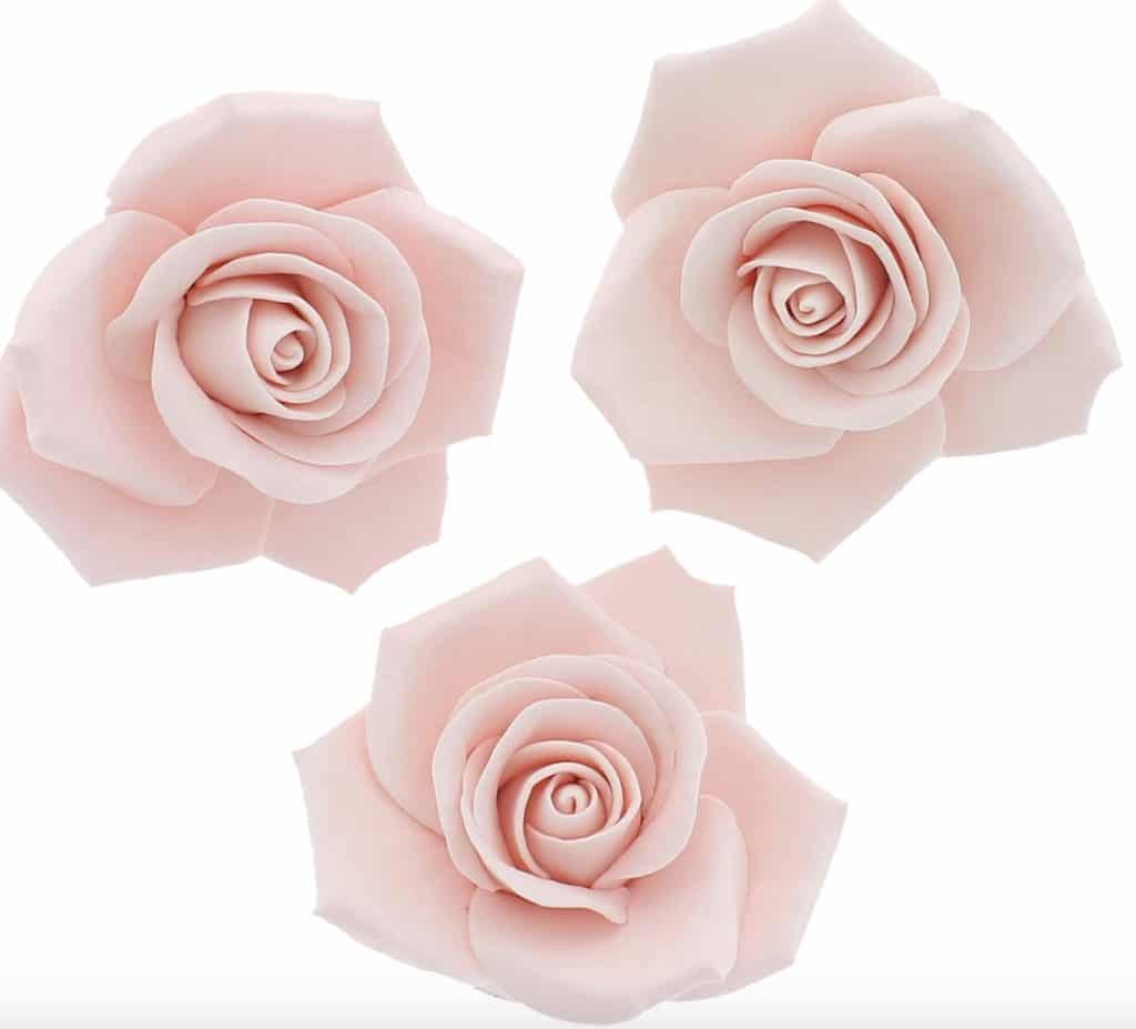 Three pale pink, intricately crafted sugar roses are arranged against a white background. These exquisite creations evoke the artistry of gourmet pink desserts with their delicate petals spiraling inward, showcasing realistic detail and texture.